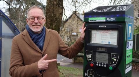 Councillor Paul Hodgkinson stands beside a parking meter outdoors. He has short brown and grey hair, a short grey beard and wears brown spectacles, a brown buttoned coat and a blue scarf. He is smiling at the camera with his left hand resting on the machine and his right hand pointing towards its screen.