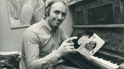 A black and white image of a man sitting in front of a piano. He holds a small record in his hands and its sleeve. He has dark hair that has been slicked over one side of his head, and is wearing a shirt. He is smiling at the camera. 