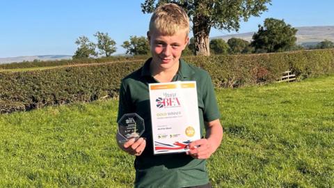 Archie Steel holding his octagonal glass award and certificate. He is standing in a field during a bright sunny day. 