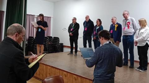 Political candidates gathered on a stage as results are read out. Each candidate is wearing a rosette. In the foreground a man is writing on a clipboard and another man is holding open a laptop.