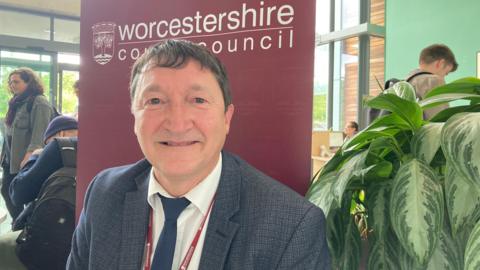 Mark Fitton, a Worcestershire County Council director, is sat down at a table with a large red banner behind him, which has the council's logo on it.