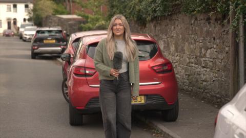 BBC reporter Holly Harrison standing in front of a red car while holding a mic