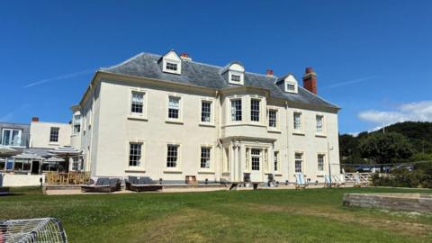 A large three storey white building, with a grey sloping roof and a dozen or so windows facing away from a blue sky and fronted by a green lawn