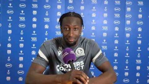Moussa Baradji is sitting with a BBC microphone infront of him with his arms leaning on a desk with a sponsor background behind him. He is wearing a Blackburn Rovers grey training top with the number 24 on it.