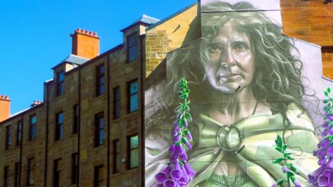 A mural of an older woman with long flowing grey hair, wearing a green shawl with a large brooch. She is surrounded by purple flowers. The mural is painted on the gable end of a sandstone tenement building.