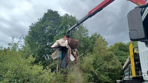 A crane carrying a load of rubbish to the back of a truck in woodland