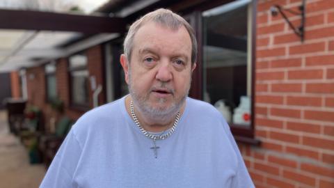 A man with grey hair and beard, he is wearing a white t-shirt and a thick silver chain with a cross on it. 