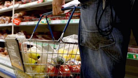 A close up of a wire supermarket shopping basket with blue handles in a supermarket. The basket has yellow, red  and green vegetables and fruit in it. The basket is held by a man, with a piece of paper in his left hand. His left jeans-clad leg is on the right. Beyond him are shelves with plastic wrapped meat. 