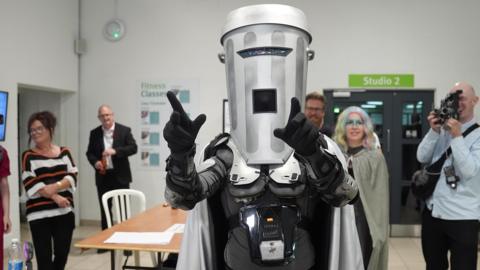 Count Binface arrives at the Clacton by-election vote count as people watch. He is pointing dramatically ahead of himself.