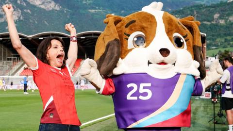 A commentator cheers and poses with UEFA Women's Euro 2025 mascot Maddli, a dog like mascot, in front of a stadium