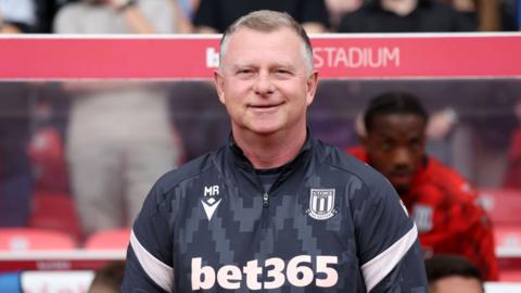 mark robins looks ahead smiling. he wears a black quarter zip top with stoke city branding