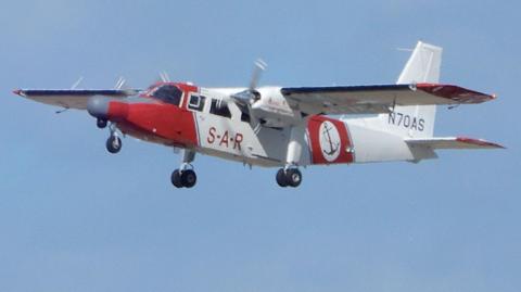 The red and white Channel Islands Air Search plane flying in daytime. It has SAR printed on its side and a logo incorporating an anchor. On its tail fin there is number N70AS. Its wheel are down.