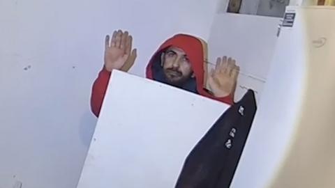 A man in a red hooded top with his hands in the air, emerging from a cupboard