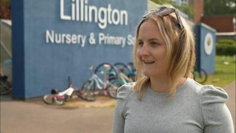 A woman outside a primary school, with bikes propped against a wall, is wearing a grey blouse. She has sunglasses resting on the top of her head and is smiling as she talks to a reporter out of view on the left hand side of the image.