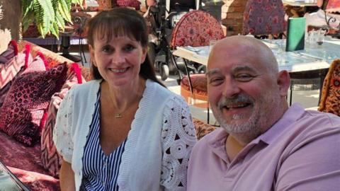 Sue and Dave Cook are sitting in an outdoor area at a restaurant. Sue has long brown hair and is wearing a blue-and-white striped top and a white cardigan. Dave is bald and has a grey beard and is wearing a pink polo shirt. They are both smiling for the camera.