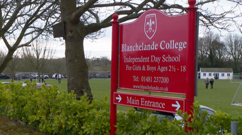 A wide shot of the college's signage. It reads "Blanchelande College, Independent Day School for girls and boys". The sign is red with white writing. A school playing field can be seen behind.