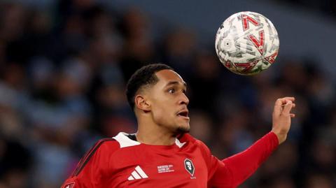 Haji Mnoga playing for Salford City, with a ball having bounced up above his head