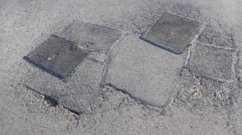 A road with six pothole repair patches next to each other, of varying age and decay