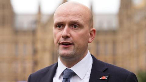 Stephen Flynn, a bald man wearing a dark jacket and white shirt, in a close-up shot with his mouth open in front of the Westminster Parliament 