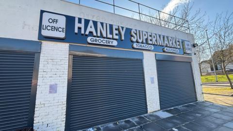 A low-roofed white brick building with shutters down. A sign on the front reads: "Hanley Supermarket - off-licence - grocery - tobacco - alcohol".