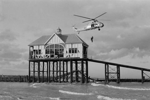 A black and white photograph shows an RAF rescue helicopter hovering above the gentle waves of the sea. A person hangs below the helicopter on a winch. In the background, the damaged remains of the RNLI boathouse can be seen.