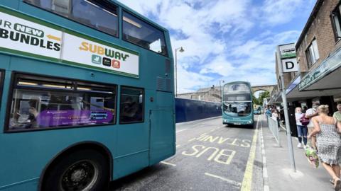 Blue double decker bus pulling away from a bus stop as another pulls in behind