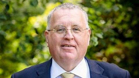 A man with short grey hair and glasses, wearing a dark suit and white tie, looks at the camera in a close-up shot 