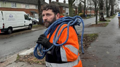 Craig Denny carries a blue rope over his shoulder as he looks to his left down a road. He is wearing orange hi-vis overalls and black gloves. He has dark curly hair and a dark beard. There are trees lining both sides of the road. On the other side of the road is a white Ipswich Borough Council van.