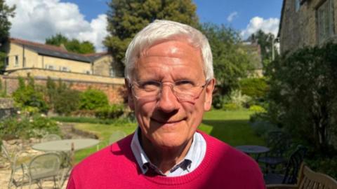 Colin Fletcher smiles for a photo taken outside his house. He has neat white hair and glasses. He is wearing a white collared shirt with a red round-necked pullover on the top. His lawn and garden furniture can be seen in the background.