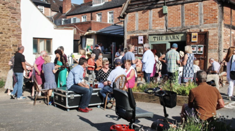 A number of people, some stood, some sat, outside a brick and timber building in the sunshine. The building's doors are open, and a sign above them reads The Barn