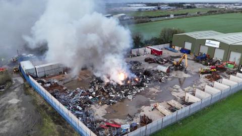 The image is a still from drone footage above the site of the fire. There is a blaze with a significant amount of grey smoke in the centre of a large, fenced yard. It's surround by a lot of scrap metal and machinery. Outside, the yard is surround by green fields. 