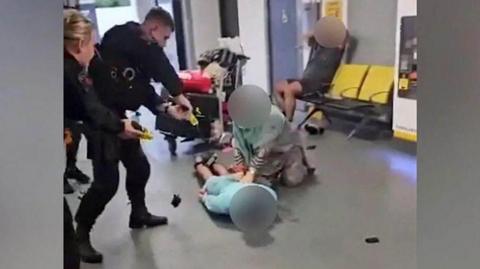 A screenshot of the video where a GMP officer kicks the man on the head at Manchester Airport, showing two police officers holding tasers pointing at a man face down on the floor, while an older woman has her hands on his back. In the background a man sits with his hands on his head watching