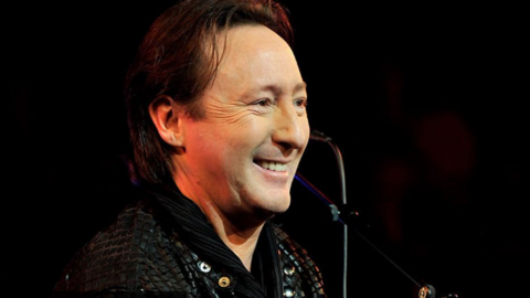 Julian Lennon smiles as he performs on stage in a black jacket