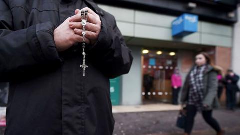 An anonymous man holds rosary beads outside an abortion clinic, with the clinic entrance in the background and an out-of-focus passerby walking in front of it