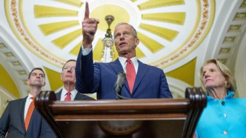 US Senate Majority Leader John Thune stands at a podium in Congress. Three senators stand behind him.