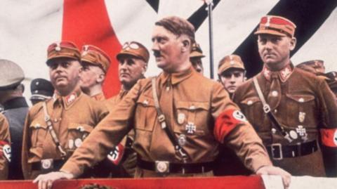 A photohraph of Hitler in a brown uniform with other men standing around him.