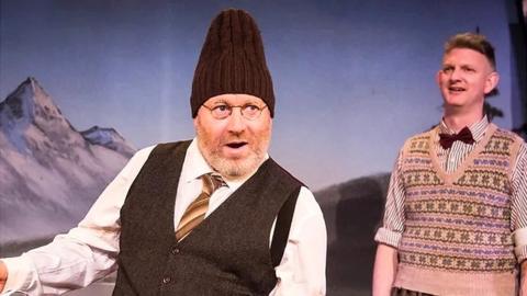 Howard Coggins on stage wearing round glasses, with a beard, white shirt and a brown tie, waistcoat and a woolly hat on.