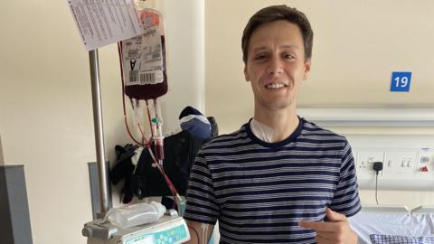 Josh is stood in hospital during a blood transfusion - he's still smiling.