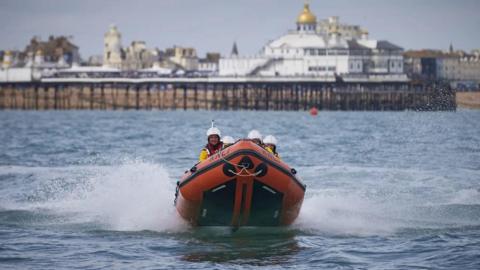 An orange lifeboat dinghy with people in it on the water. A pier with a gold roof is in the background.