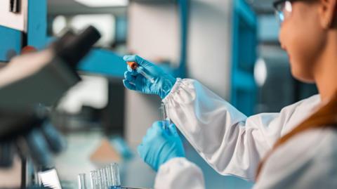 A woman with blue gloves on and white lab suit holding a lab beaker and a pipette. The photo is taken from behind her shoulder.