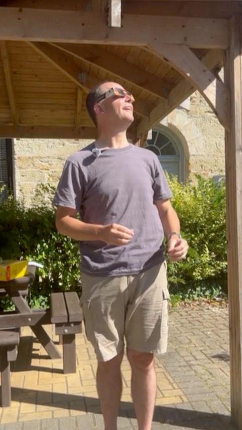 A man under a wooden canopy with a picnic bench and bush behind him. He is wearing a top and shorts and eclipse glasses and stating to the right.
