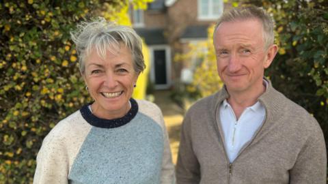 Liz and Andrew Potter look into camera with their front door in the background. Liz has short white hair and is wearing a turquoise and white jumper. Andrew has short white hair and is wearing a grey cardigan and white shirt.