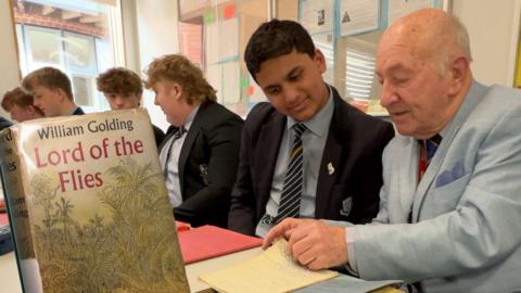 Mick (right) sits with current pupils of the school. Mick is wearing a light blue suit and a tie, and is looking through an old text book with a pupil who is smiling. Other pupils, all wearing black blazers, blue shirts and blue striped ties, are in the background. In the foreground there is a copy of Lord of the Flies, with a jungle scene on the cover. 