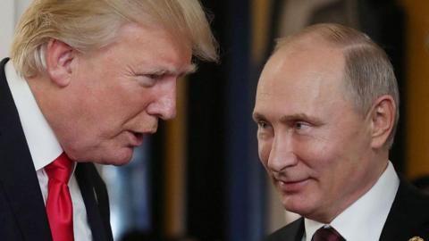 A close of up Putin looking left at Trump as he speaks to him at the Asia-Pacific Economic Cooperation (APEC) leaders' summit in 2017. Trump is wearing a red tie, and both leaders are pictured in front of a blurred background. 