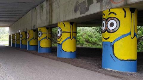 A row of eight painted stanchions under a motorway bridge, looking like yellow minions wearing blue dungarees and holding up the bridge