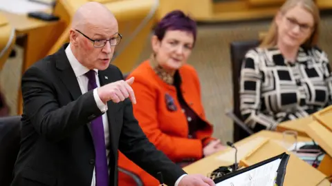 First Minister John Swinney stands in the chamber pointing downwards during FMQs. Justice Secretary Angela Constance and Social Security Secretary Shirley-Anne Somerville are in the background seated.