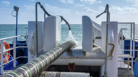 A black and green striped thick interconnector cable going off the back of a ship into the sea. There is a hose on top of the cable spraying water on it. 