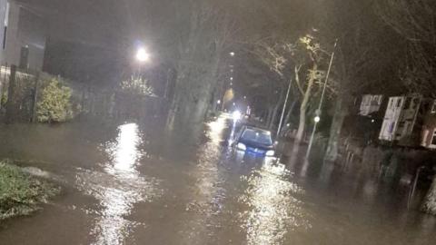 A severely flooded street at night. A car is submerged up to its headlights in the standing water.