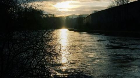 The River Leven photographed near sunset. The banks are in shadow with branches on one side and buildings visible on the other
