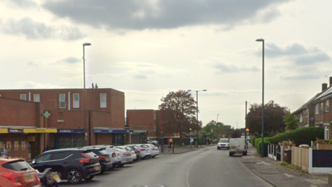 A parade of shops by a road. Cars and vans are parked in the highway